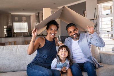 zas Portrait of smiling family sitting on couch holding cardboard roof and looking at camera. African and indian parents with funny daughter holding cardboard roof over heads while sitting on sofa in their new home. Happy mother and father with daughter in new house at moving day with copy space.